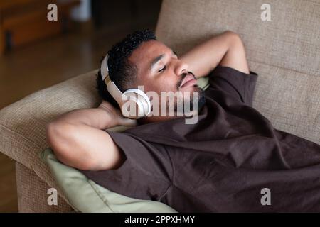 From above relaxed ethnic African American male in casual t shirt lying on comfortable sofa with closed eyes while listening to music on wireless head Stock Photo