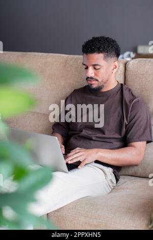 Concentrated young ethnic African American male freelancer in casual outfit typing information on laptop keyboard while working remotely from home and Stock Photo