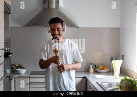 Positive young ethnic African American male in casual outfit smiling and singing in wooden spatula while listening to music in wireless headphones wit Stock Photo