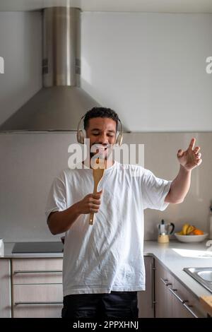 Positive young ethnic African American male in casual outfit smiling and singing in wooden spatula while listening to music in wireless headphones wit Stock Photo