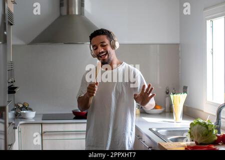 Positive young ethnic African American male in casual outfit smiling and singing in wooden spatula while listening to music in wireless headphones wit Stock Photo