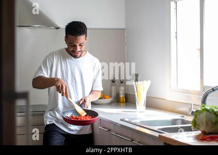 Positive young ethnic African American male in casual clothes preparing delicious dish while frying tomatoes on pan and mixing ingredients with wooden Stock Photo