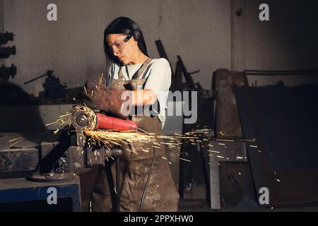 Concentrated young woman in forge apron and gloves wearing protective eyeglasses and cutting iron detail with angle grinder in flying sparkles while w Stock Photo