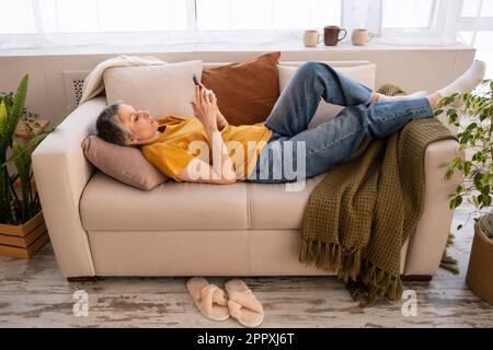 Relaxed mature woman surviving cancer with silver hair in casual clothes resting on sofa in modern living room decorated with fresh green flowers whil Stock Photo