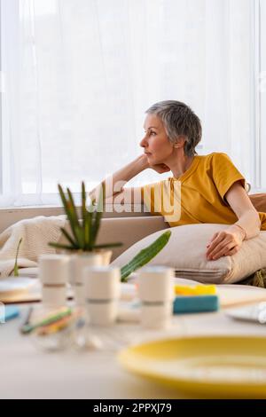 Mature woman recovering from cancer in Perm Russia relaxing on couch in cozy living room with wooden table decorated with green cactus flower pots and Stock Photo