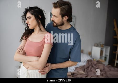 Bearded man calming displeased girlfriend in pajama in bedroom,stock image Stock Photo