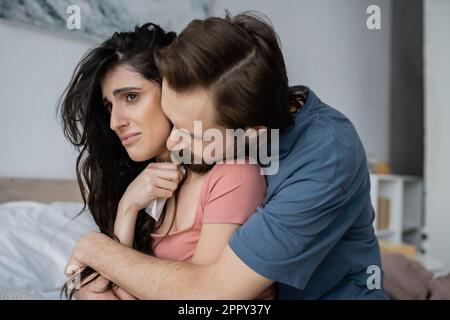 Bearded man hugging sad girlfriend with napkin in blurred bedroom,stock image Stock Photo