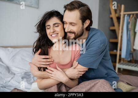 Overjoyed man hugging brunette girlfriend in pajama on bed at home,stock image Stock Photo