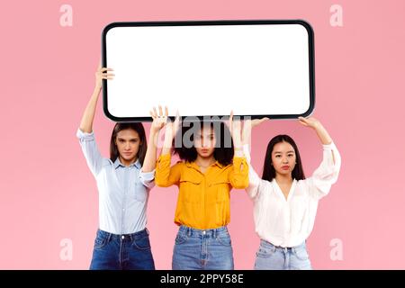 Three multiracial ladies holding huge smartphone with white empty screen on pink studio background, mockup Stock Photo