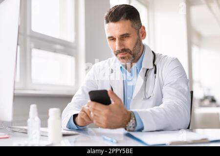 Serious male doctor in clinic wearing white coat uniform using cellphone, sitting at desk, consulting patients online Stock Photo