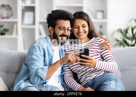 Relaxed multicultural couple sitting on sofa at home, using smartphone Stock Photo