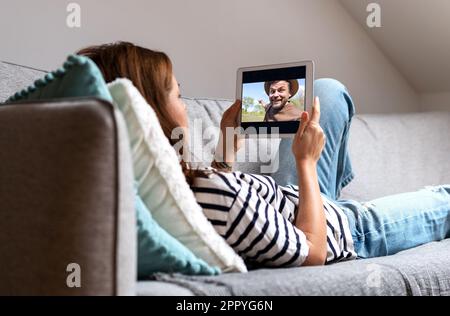 Woman lying down on sofa at home watching travel video blog in internet using her digital tablet. Stock Photo