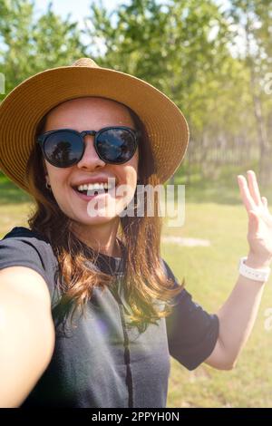 Woman wearing sunglasses and straw hat waving hand and taking a selfie while walking outdoors. Stock Photo
