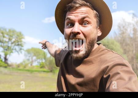 Travel video blogger wearing straw hat recording video in wild nature, looking at camera with contented emotions and pointing in the distance. Stock Photo