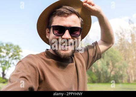 Man wearing dark glasses and straw hat taking a selfie outdoors in summer, mental wellbeing and positive emotions concept. Stock Photo