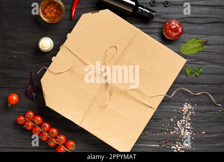 Pizza in a closed cardboard box, sauces, olive oil with spices, fresh vegetables on a dark wooden background top view. Food delivery. Stock Photo