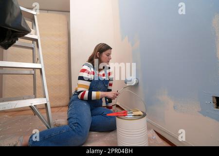 Kneeling woman dressed in overalls and striped blouse, profile view, painting a white wall with a roller with blue paint, inside an empty room with a Stock Photo