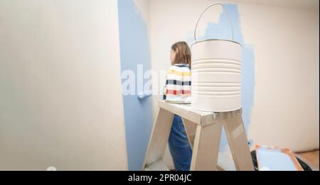 Close-up of a jar of paint on a ladder with a background of a standing woman dressed in overalls and striped blouse, seen from behind, painting a whit Stock Photo