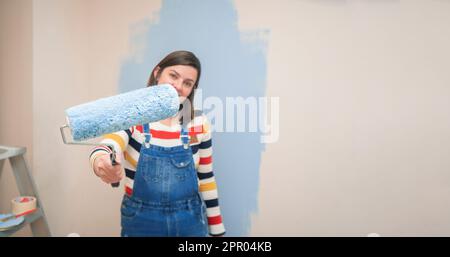 Close-up of roller with blue paint in the hand of a standing woman dressed in overalls and striped blouse, seen from the front, against background of Stock Photo