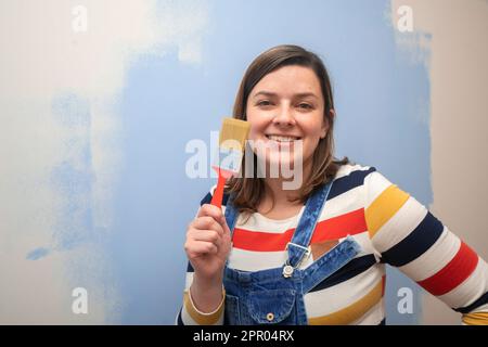 Beautiful woman standing dressed in overalls and striped blouse, smiling at the camera with a brush in her hand against a background of a white wall h Stock Photo