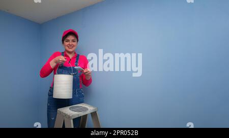 Beautiful woman dressed in overalls and red t-shirt standing on a metal ladder, seen from the front, holding the brush and the paint jar next to a blu Stock Photo