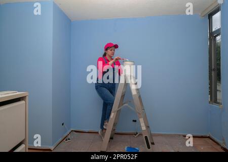 Beautiful woman dressed in overalls and red t-shirt going up a metal ladder with a jar of paint next to a blue wall in an empty room Stock Photo