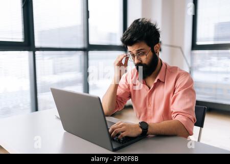 Thoughtful Indian businessman wearing casual clothes working on laptop sitting at table in light office room on background of window. Stock Photo