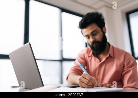 Selective focus portrait of Indian businessman using laptop analyzing marketing report, writing notes siting at table by window. Stock Photo