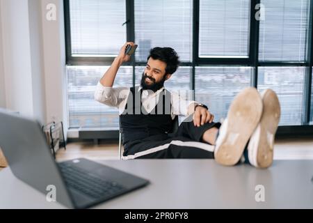Portrait of happy excited businessman sitting at office desk with feet up on table with laptop, finished work, job done, completed all tasks Stock Photo