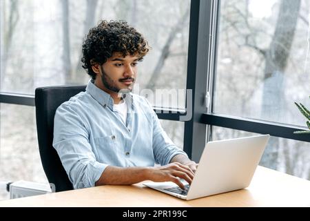 Smart successful confident indian or arabian man, company seo, IT specialist, HR manager, works on a laptop while sitting at a work desk in the office, looks at the laptop screen, smile Stock Photo