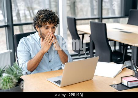 Shocked amazed indian or arabian businessman, company employee, ceo, manager, sits at workplace in the office, looks in surprised at a laptop screen, reads unexpected news, rejoices in success Stock Photo