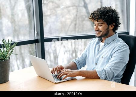 Positive indian or arabian businessman, company employee, ceo, manager, sits at workplace in the office, working on a laptop, planning new project, developing strategy and promotion Stock Photo