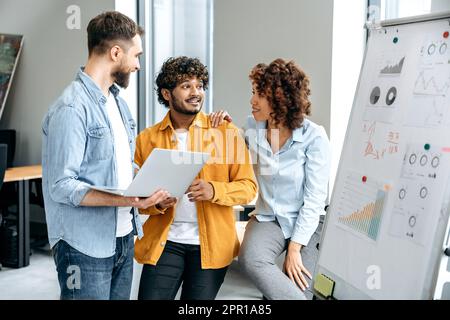 Positive busy successful colleagues of different nationalities, office employees, managers, stand in a modern office near the marker board, analyze and discuss financial charts, profit, risks Stock Photo