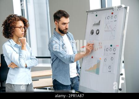Focused busy successful colleagues of different nationalities, a man and a woman, office employees, stand in a modern office near the marker board, concentrated analyze financial charts, profit, risks Stock Photo
