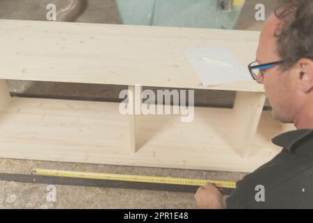 Carpenter takes measurements of a piece of furniture with a tape measure in his craftsman's workshop Stock Photo