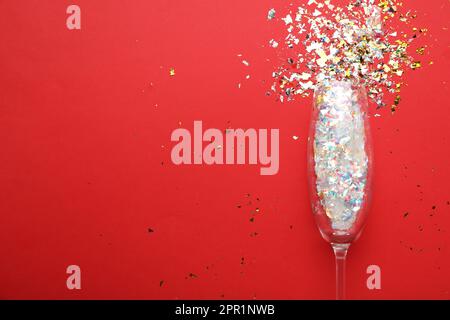 Shiny confetti spilled out of champagne glass on red background, top view. Space for text Stock Photo