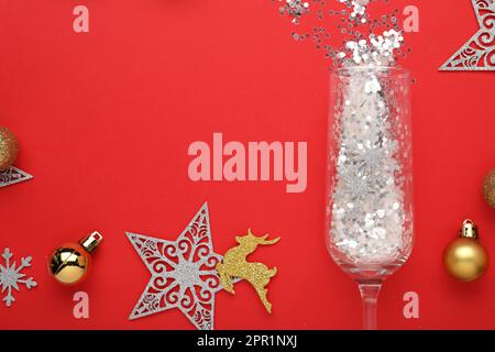 Flat lay composition with shiny confetti spilled out of champagne glass near Christmas decorations on red background, space for text Stock Photo