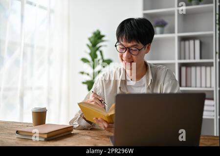 Portrait of young Asian man writing on notebook while working with laptop computer in the living room. Stock Photo
