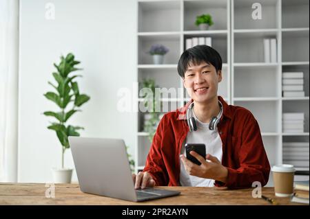 Cheerful young college student using laptop computer and smartphone while study in the study room. Stock Photo