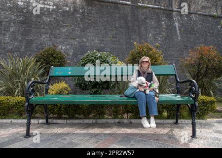 Kotor, Montenegro, Apr 9, 2023: A woman with a dog looking tiny while sitting on an oversized bench Stock Photo