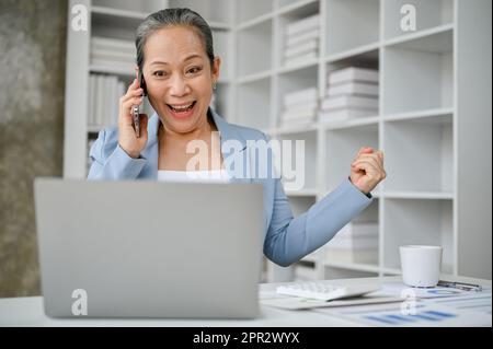 Happy and overjoyed senior businesswoman feeling exited after hearing a good news from the phone  call at work. Stock Photo