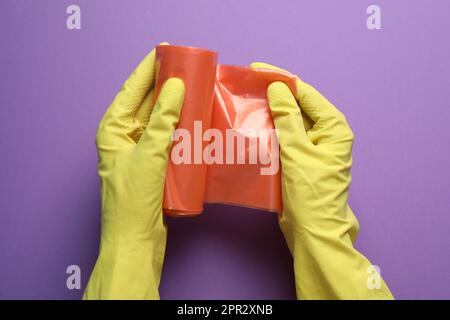 Janitor in rubber gloves holding roll of orange garbage bags over purple background, top view Stock Photo