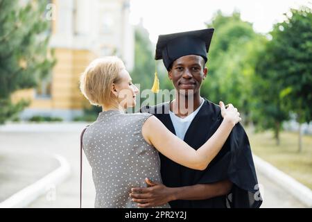 Happy afro american student in graduation mantle and hat thankfully hugging attractive middle aged woman. Higher education, graduate from university Stock Photo