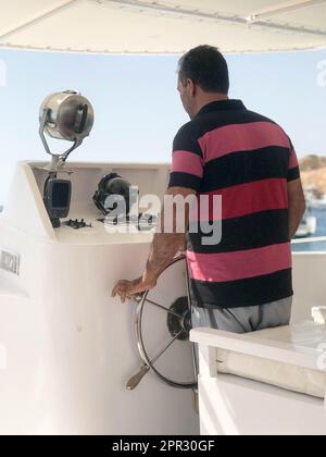 The swarthy captain of the man in the red striped T-shirt manages the boat, the boat at the helm. Stock Photo