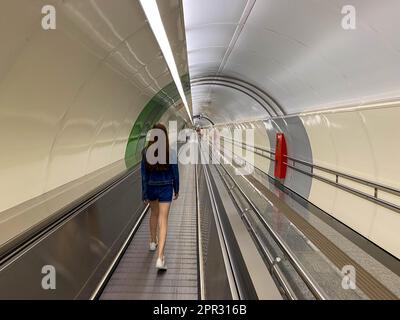 A beautiful girl walks along the travolator in the subway tunnel to go to another underground station. View from the back. Stock Photo