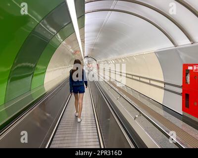 A beautiful girl walks along the travolator in the subway tunnel to go to another underground station. View from the back. Stock Photo