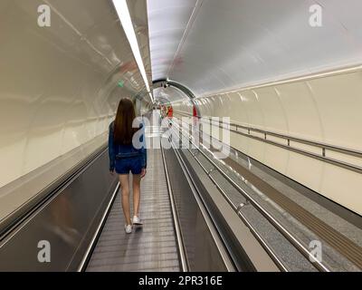 A beautiful girl walks along the travolator in the subway tunnel to go to another underground station. View from the back. Stock Photo