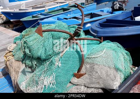 Old rusty anchor on top of a pile of fishing nets with fishing boats in the background Stock Photo