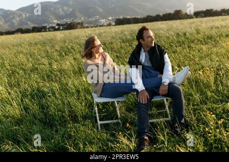 Young happy couple sitting on chairs in feather grass field on summer sunset day. Stock Photo