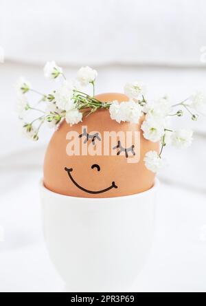 Easter egg with cute face in floral wreath crowns in egg cup on white background. Easter egg with flowers and sleepy eyes in sunny light. Happy Easter Stock Photo